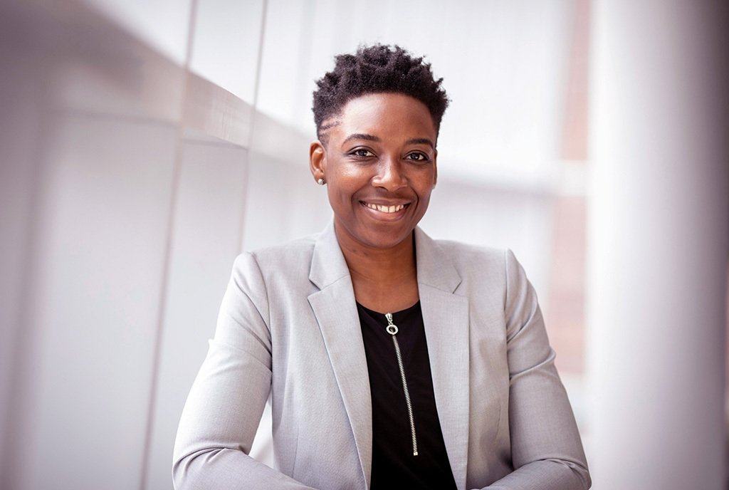 A portrait of a smiling Black woman executive, wearing a gray suit jacket and a black blouse. She is standing in a well-lit hallway.