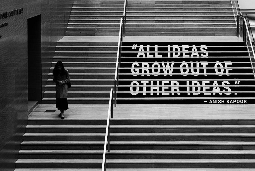 A woman walking down a staircase that has the phrase “All ideas grow out of other ideas. - Anish Kapoor” plastered onto the stair risers.